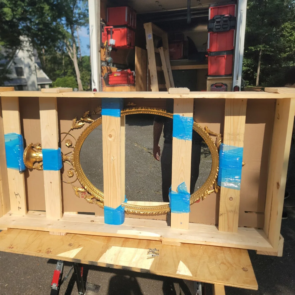 Ornate gilded mirror braced inside a custom-built wooden crate on sawhorses beside an open work truck